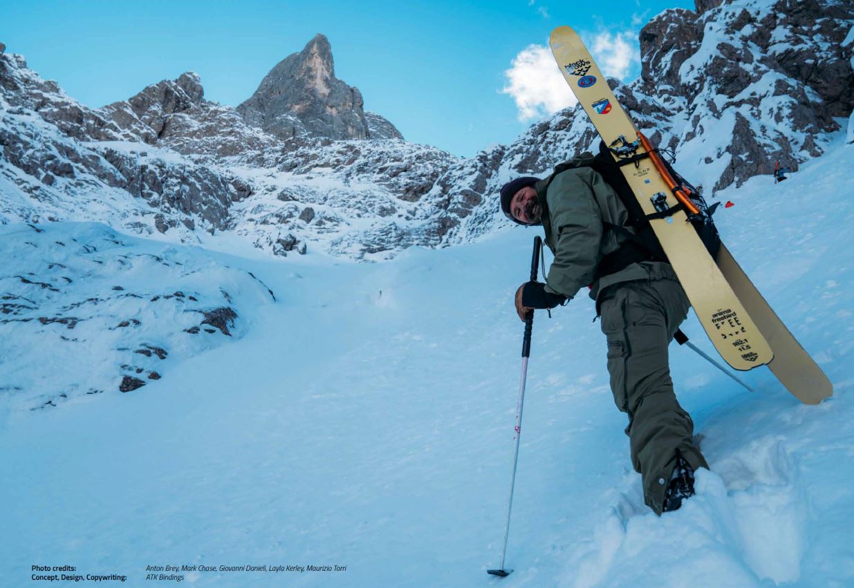 Ein Skifahrer mit gelber Skiausrüstung und breitem Lächeln steigt eine verschneite Bergflanke mit Skiern auf dem Rücken empor, umgeben von felsigem, schneebedecktem Gelände.